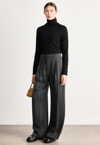 Camisola de gola alta preta, calças largas cinzentas de cintura alta, combinadas com sapatos pretos de plataforma e uma pequena mala bege.