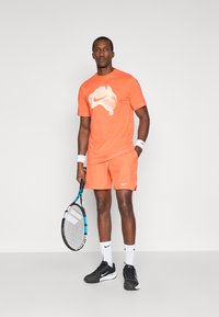 Πορτοκαλί αθλητική μπλούζα με λευκό γραφικό, πορτοκαλί σορτς που ταιριάζουν, μαύρα παπούτσια και ρακέτα τένις με μπλε λεπτομέρεια.