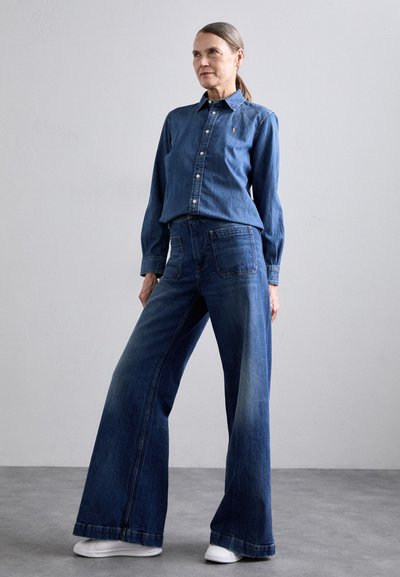 Denim shirt with button-down collar, long sleeves, and logo detail, paired with wide-leg blue denim jeans. White sneakers complete the outfit.
