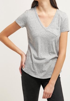 Frau trägt ein hellgraues V-Ausschnitt-T-Shirt mit kurzen Ärmeln und einer kleinen Brusttasche, kombiniert mit schwarzen Hosen, steht mit einer Hand an der Hüfte.