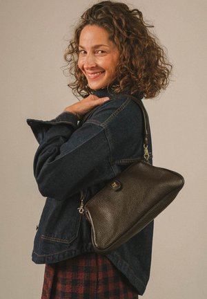 Femme aux cheveux bouclés souriante, portant une veste en jean foncé et un sac à épaule marron texturé sur un fond neutre.