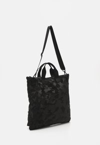 Czarna torba tote z nylonu w kamuflażu, z podwójnymi uchwytami, odpinanym paskiem i zapięciem na zamek. Płaski, prostokątny kształt o gładkiej teksturze.