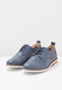 Scarpe oxford in pelle blu con tomaia testurizzata, lacci sottili e suole bianche a contrasto. Presentano un piccolo dettaglio marrone sul tallone.
