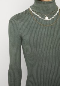 Femme portant un pull col roulé côtelé vert avec des colliers superposés en or et perles blanches à motifs floraux sur un fond uni.