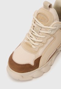 Bež tenisice izrađene od mrežastog i antilop materijala, s debelim đonom, vezicama i logom na jeziku. Okruženi prst i teksturirani detalji.