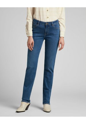 Woman wearing fitted blue jeans, cream-colored button-up shirt, and white heeled ankle boots, standing against plain background.