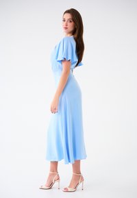 Hellblaues Midi-Kleid mit schwingenden Ärmeln, tailliertem Schnitt, fließendem Rock, glatter Textur und kombiniert mit nude-farbenen High Heels.