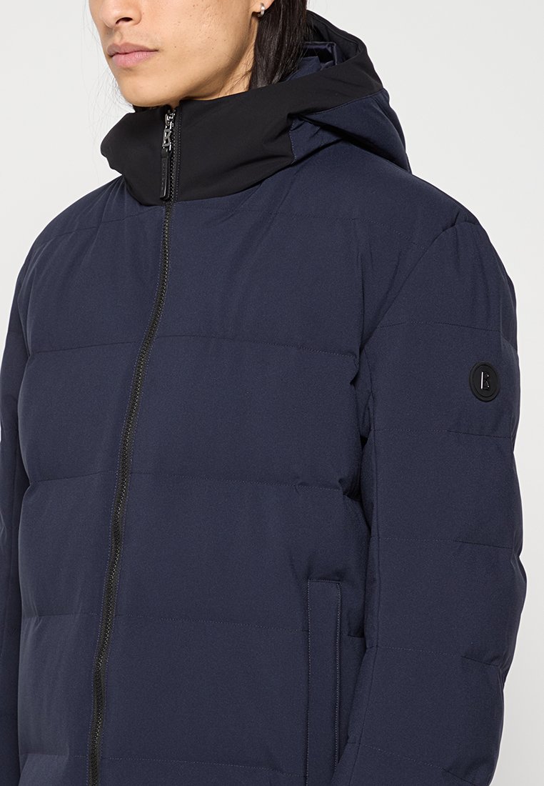 Marineblaue Steppjacke mit Reißverschluss, hohem Kragen und schwarzer Kapuze. Verfügt über horizontale Nähte und ein rundes Logo am Ärmel.
