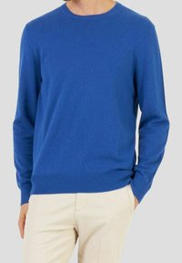 Man som bär en blå långärmad crewneck-tröja och ljusbeige byxor med en hand i fickan mot en neutral bakgrund.