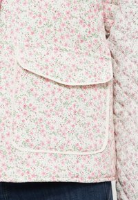 Ύφασμα με καπιτονέ ραφή και ροζ λουλουδάτο μοτίβο σε ένδυμα με μπροστινή τσεπάκι με καπάκι, φορεμένο πάνω από σκούρο μπλε τζιν.