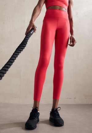 Leggings taille haute, ajustés, de couleur rouge vif avec une texture lisse, associés à des chaussures de sport noires. Personne tenant une corde noire texturée.