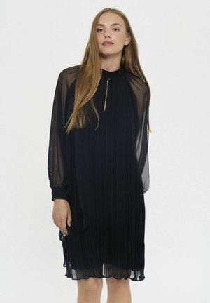 Frau mit langen blonden Haaren, die ein knielanges schwarzes plissee Kleid mit transparenten langen Ärmeln trägt, steht vor einem schlichten hellen Hintergrund.