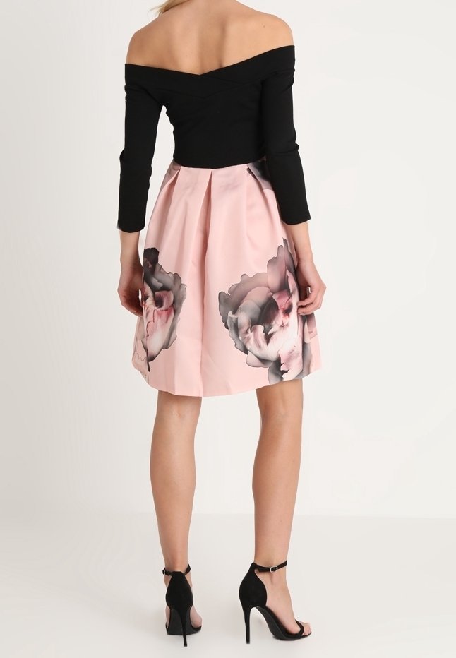 Svart topp med avskuren axel och långa ärmar, i kombination med en rosa kjol med blommönster och svarta högklackade sandaler.