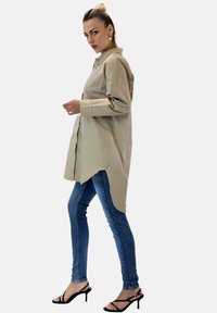 Ljus beige lång skjorta med knäppning, rundad nederkant och långa ärmar, tillsammans med blå jeans och svarta klacksandaler.