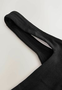 Gros plan sur une sangle en tissu noir avec des coutures visibles sur un fond clair uni, probablement partie d'un vêtement ou d'un accessoire.