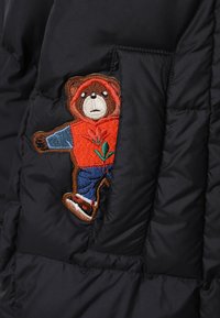 Juodas pūkuotas švarkas su bear siuvinėta raudonoje džemperyje, mėlynos kelnės ir raudoni batai, esantys kairėje šoninės kišenės pusėje.