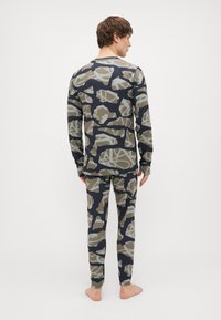 Ilgų rankovių marškiniai ir kelnių komplektas tamsiai mėlynos kamufliažo rašto. Audinys atrodo minkštas, su pilkomis ir smėlinėmis detalėmis, turi apvalią kaklo iškirptę ir prigludusį siluetą.