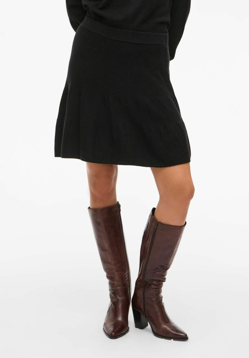 Bottes en cuir marron montantes jusqu'au genou avec un bout pointu et un talon épais, dotées d'une fermeture éclair sur le côté et d'une texture lisse. Portées avec une jupe noire.