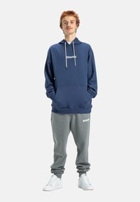 Námornícky modrá mikina s bielym logom, sťahovacími šnúrkami a prednou vreckom, skombinovaná so šedými teplákmi s logom, doplnená bielymi teniskami.