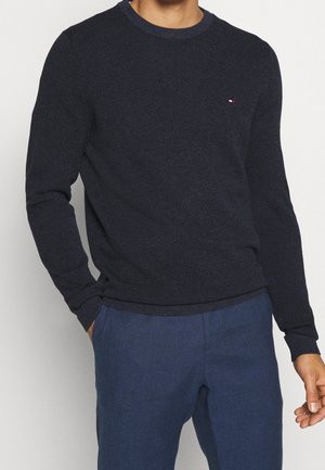 Pull en maille marine avec un col rond, des poignets et un ourlet côtelés. Présente un petit logo au niveau de la poitrine. Associe avec un pantalon bleu.