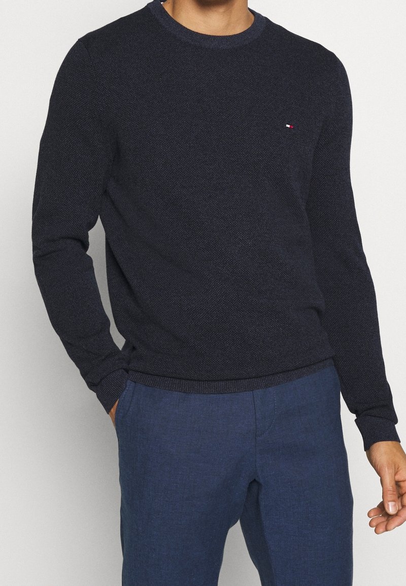 Laivastonsininen neulepusero, jossa on pyöreä kaula-aukko, röyhelömuotoiset hihansuut ja helma. Rinnassa on pieni logo. Yhdistetty sinisiin housuihin.