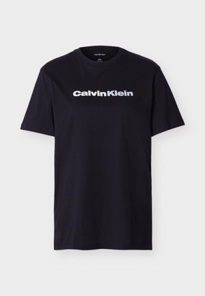 Czarna koszulka z krótkim rękawem i okrągłym dekoltem z białym logo "Calvin Klein" na środku klatki piersiowej, przedstawiona na jednolitym jasnym tle.