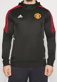 Veste de sport noire avec accents rouges, col zippé, manches longues, arborant le logo de Manchester United et le branding Adidas sur la poitrine.