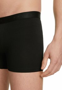 Czarne bawełniane bokserki z gładką fakturą i elastycznym pasem, z wytłoczonym logotypem. Zbliżenie na nogę i rękę.