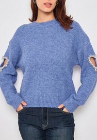 Pull tricot bleu clair à manches longues, présentant des découpes au niveau des coudes embellies de accents décoratifs. Tissu texturé.