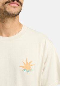 Cr�ème-kleurig T-shirt met een oranje sterafbeelding en blauwe tekst "RVCA" geborduurd op de linkerborst. Zachte katoenstof.