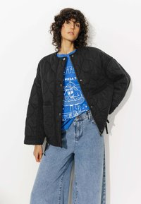 Blouson bomber noir matelassé avec col rond et boutons-pression, porté sur un t-shirt graphique bleu et associé à un jean en denim taille haute.