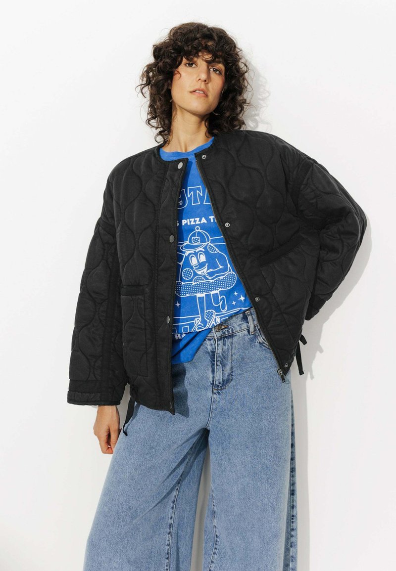 Blouson bomber noir matelassé avec col rond et boutons-pression, porté sur un t-shirt graphique bleu et associé à un jean en denim taille haute.
