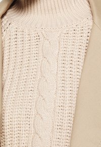 Pullover lavorato a maglia beige chiaro con colletto alto a costine e motivo centrale a treccia, accompagnato da un tessuto beige liscio su un lato.
