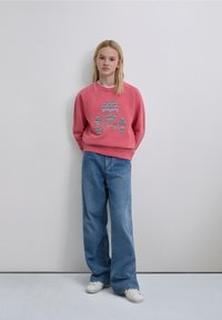 Pink sweatshirt featuring a patterned skull design, paired with loose-fitting blue jeans and white sneakers. Smooth fabric textures visible.