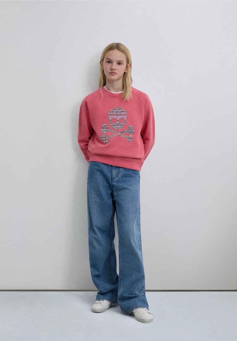 Pink sweatshirt featuring a patterned skull design, paired with loose-fitting blue jeans and white sneakers. Smooth fabric textures visible.