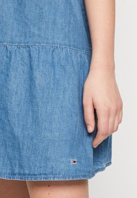Vestido de mezclilla azul claro con dobladillo rizado. Presenta mangas cortas y una pequeña etiqueta de logo cerca del dobladillo inferior. Textura suave.