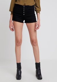 Shorts de mezclilla negra con dobladillo deshilachado, con una cintura alta, cierre frontal de cinco botones y bolsillos. Llevados con botas negras hasta el tobillo.