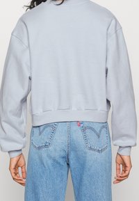 Personne portant un sweat-shirt court gris clair et un jean en denim bleu taille haute avec des poches arrière et une étiquette de marque rouge.