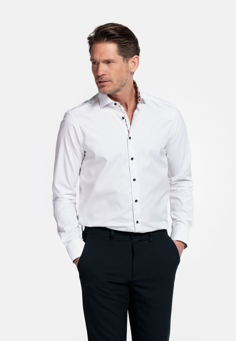 Witte knopsluitende shirt met zwarte knopen en een bloemenpatroon aan de binnenkant van de kraag, gecombineerd met donkere broek. Gladde stof en een snit op maat.