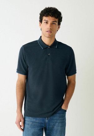 Młody mężczyzna o kręconych włosach, ubrany w ciemnogranatową koszulkę polo z wzorzystym kołnierzykiem i krawędziami rękawów, w połączeniu z niebieskimi dżinsami, stojący na białym tle.