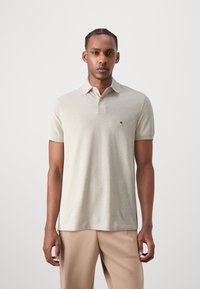 Helles beiges Poloshirt aus weichem Stoff, mit klassischem Kragen, drei Knöpfen und kleinem Logo auf der linken Brust.