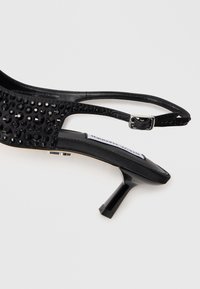 Sandale cu toc înalt, tip slingback, de culoare neagră, cu toc mic bloc, partea superioară cu ținte și curea ajustabilă, expuse pe un fundal alb.