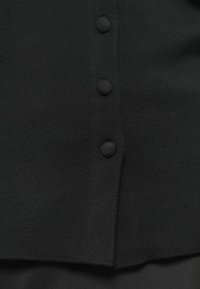 Černý pletený cardigan s rovný střih, pěti knoflíky potaženými látkou a hladkým povrchem s mírně roztrhaným lemem.