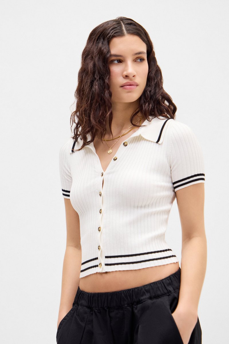 Polo scurt din tricot alb cu nervuri, cu mâneci scurte, accente cu dungi negre și nasturi aurii, având un design ajustat și o lungime scurtă.