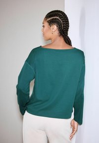 Zielona bluzka z długim rękawem o szerokim dekolcie i luźnym kroju, charakteryzująca się gładką fakturą i minimalnym przeszyciem. Nosi się ją z jasnymi spodniami.