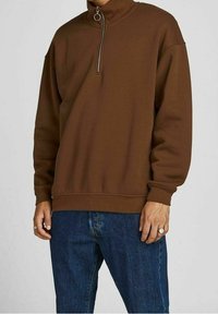 Sudadera marrón con cremallera y alto cuello, con puños acanalados, combinada con pantalones vaqueros azul denim. La tela parece suave y ligeramente texturizada.