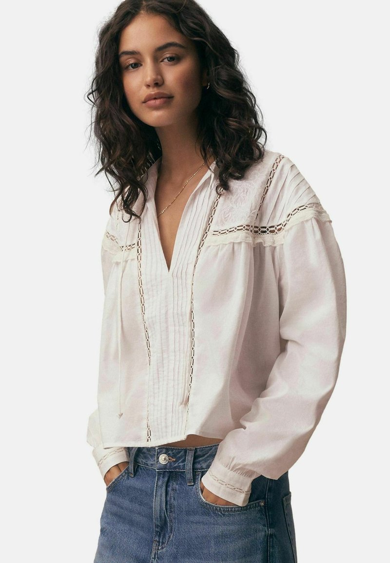 Mladi žena tamne kovrčave kose nosi bijelu bluzu s dugim rukavima i detaljima od čipke te plave traperice, ruke u džepovima.