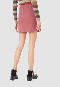 Mujer vestida con una minifalda roja, una camiseta de rayas de manga larga y botas negras hasta el tobillo con detalles de cremallera dorada, de pie contra un fondo liso.