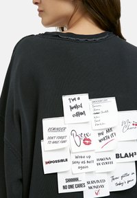 Nahaufnahme des Rückens eines schwarzen Sweatshirts mit mehreren aufgenähten weißen Stoffpatches, die motivierende Zitate und eine Einkaufsliste zeigen.