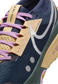 Gros plan d'une chaussure de course Nike Trail bleu marine avec des lacets lavande, des accents beige et un swoosh argenté réfléchissant ainsi que des points.
